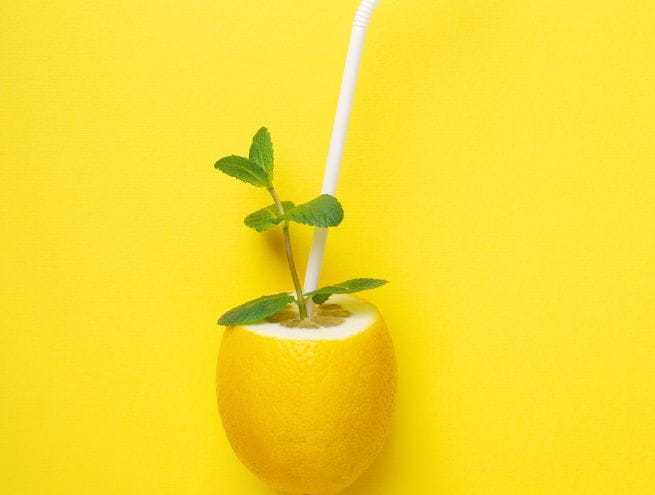 A half lemon with a straw and mint leaves on a bright yellow background, symbolizing a fresh drink.