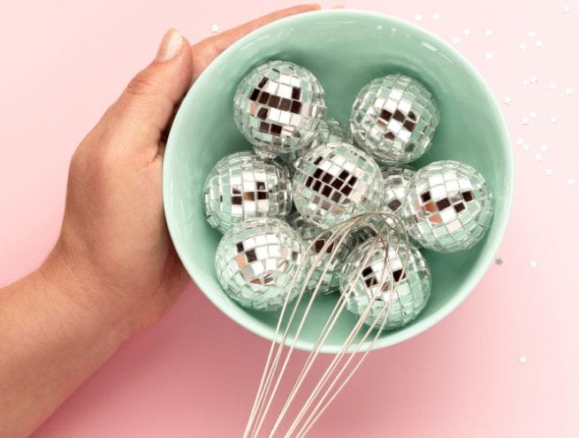 A turquoise bowl filled with small silver disco balls and a whisk, held by a hand on a pale pink background with silver stars.