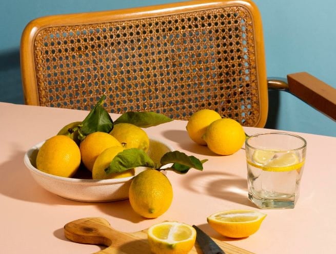 A bowl of fresh lemons and lemon slices on a table, with a glass of lemon water and a wicker chair in the background.