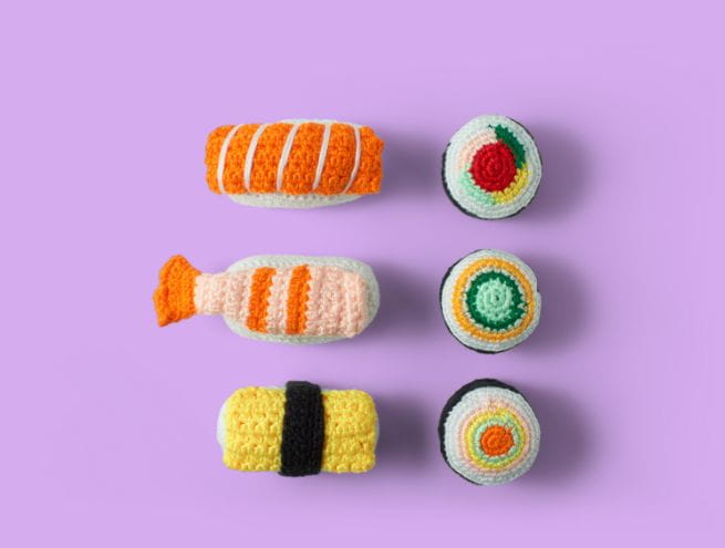 Six pièces de sushi en crochet coloré sur un fond violet, représentant différents types de sushi.