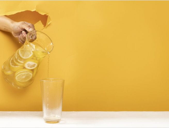 A hand holding a pitcher of lemonade, pouring into a glass, set against a bright yellow background.