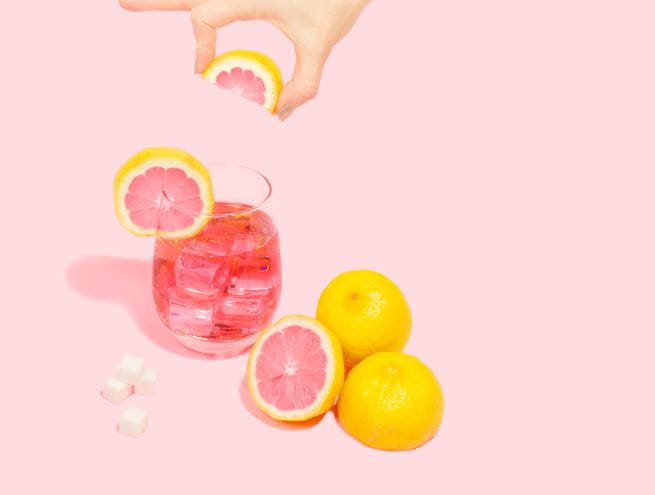 A glass filled with pink iced drink, garnished with grapefruit slices and whole lemons on a pastel pink background.