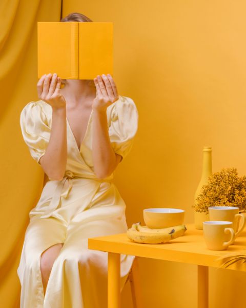 Person sitting in an entirely yellow room, dressed in a cream-colored dress, holding a yellow book in front of their face, with a table decorated with cups and a banana.