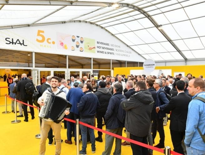 Entrée d'un hall du salon SIAL Paris avec un groupe de visiteurs attendant leur accès, un musicien jouant de l'accordéon au premier plan, et une signalétique jaune indiquant les boissons et spiritueux.