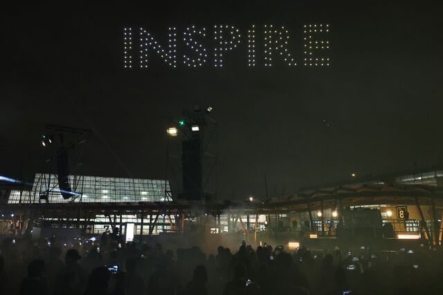 Nighttime show featuring the word "INSPIRE" displayed in the sky using drones, with a crowd of spectators holding their phones. The event takes place outside an exhibition center.