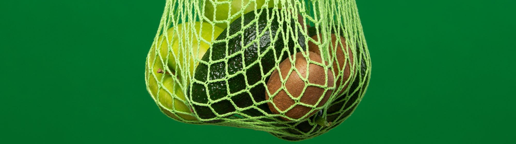 Green and brown fruits, including apples and avocados, hanging in a green mesh bag against a solid green background.