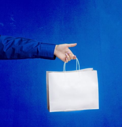 Hand holding a white shopping bag against a bright blue background, symbolizing shopping or forming new partnerships.