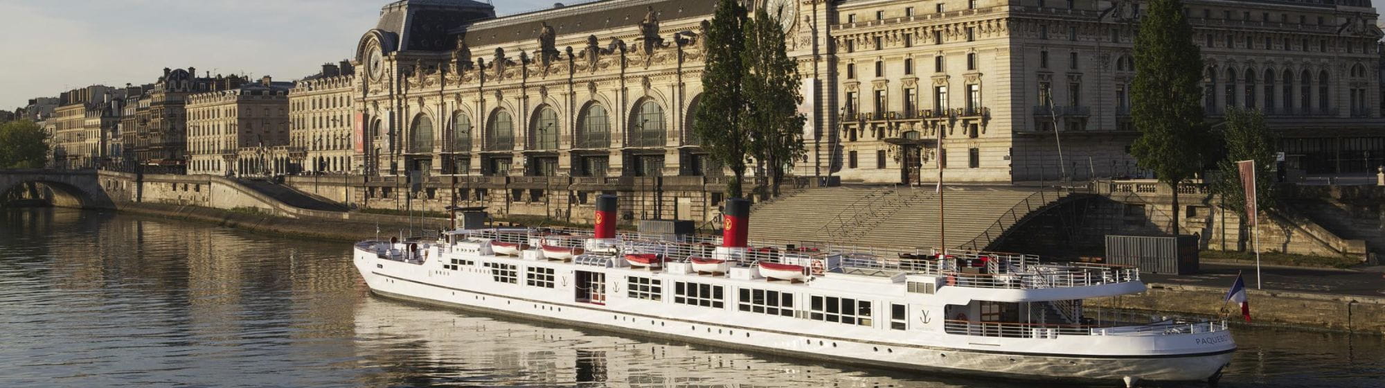 A large white cruise ship moored along the Seine in Paris, in front of the Musée d'Orsay, with red buoys and black and red chimneys, bathed in the golden light of late afternoon