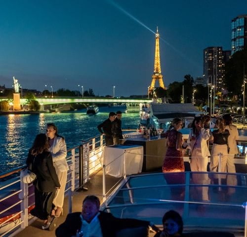 Guests relax on the deck of a cruise ship in the evening, with the Eiffel Tower and the Statue of Liberty visible in the background, illuminated against the night sky