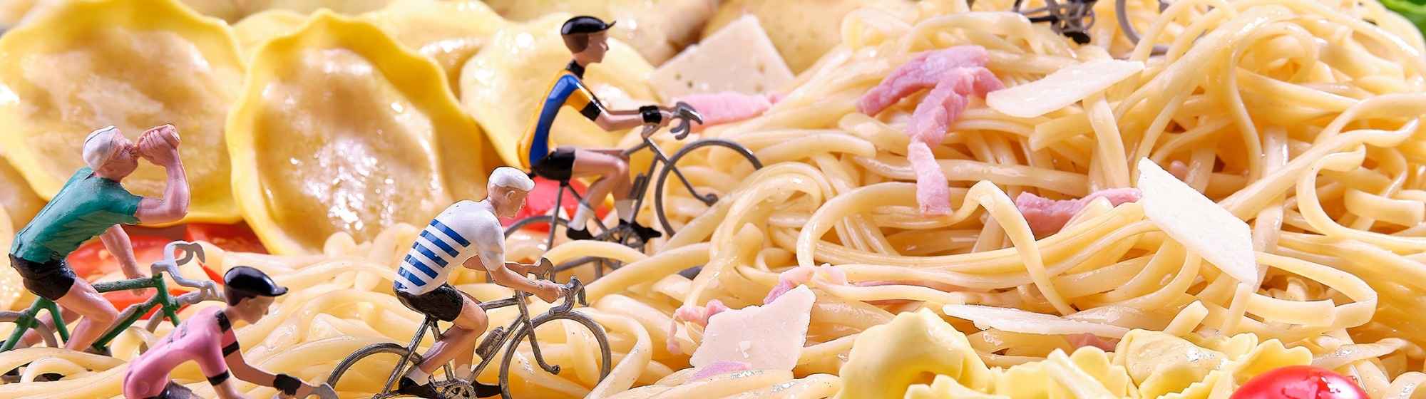 Miniatures de cyclistes pédalant sur un paysage de pâtes, comprenant des spaghettis, des gnocchis, et des tortellinis, avec des pommes de terre et de la laitue en arrière-plan.